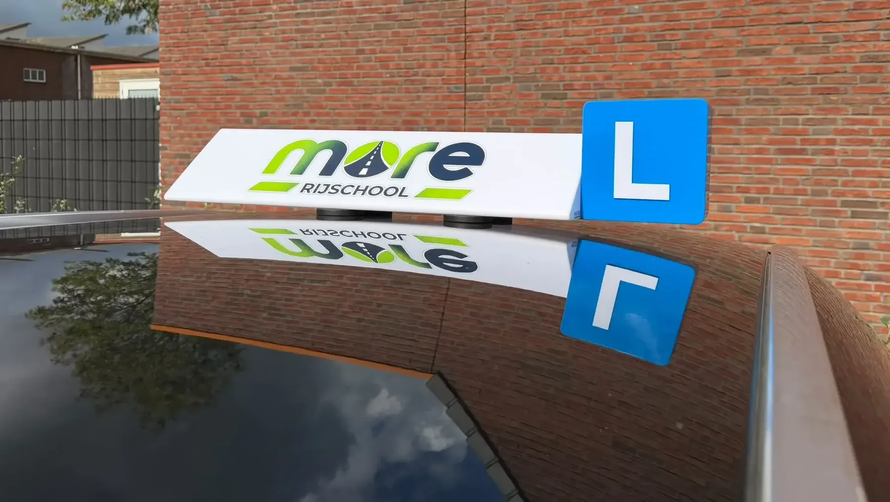 Daklicht met het logo van Rijschool More en een blauw L-bord op het dak van de lesauto